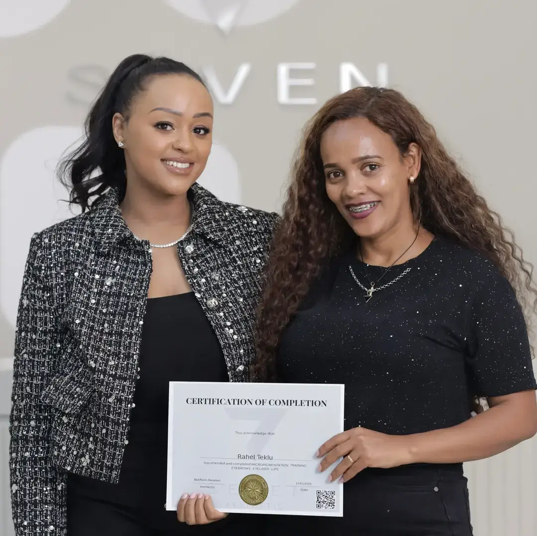 Permanent Makeup Certification at Seven Aesthetics: Teacher Betlehem Bereket with student Elsa Elias holding her certificate of completion, celebrating with champagne after completing the course.